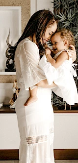 A woman in a white dress lovingly holds a small child, also in white, inside a warmly decorated room with plants and decorative elements.