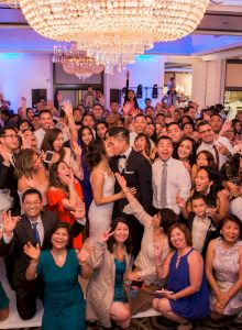 A large group at a celebration, likely a wedding, with a couple in the center surrounded by smiling guests under a chandelier.