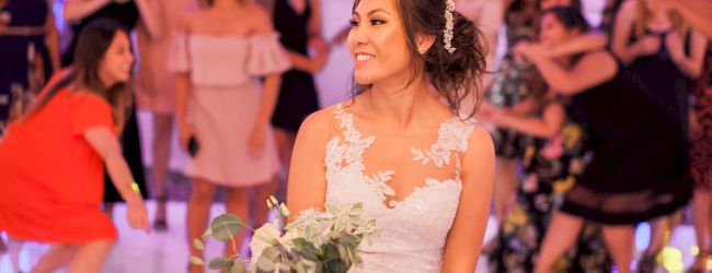 A bride in a wedding dress holds a bouquet, preparing for the bouquet toss. Guests in dresses gather behind her in a decorated venue.