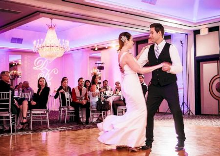 A couple is dancing at a formal event in a decorated ballroom with guests seated around, under a large chandelier.