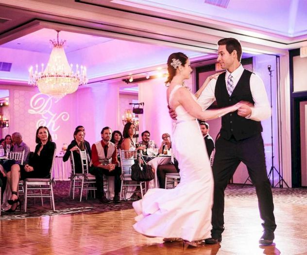 A couple in wedding attire dances at a banquet hall as guests watch under chandeliers and purple lighting.