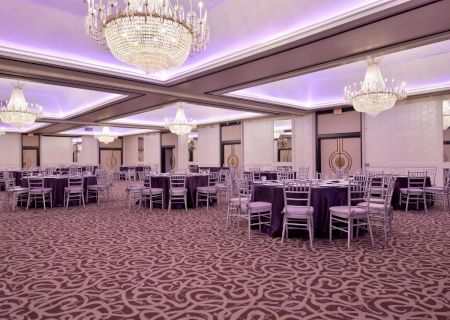 An elegantly decorated ballroom with chandeliers, tables, and chairs, featuring a patterned carpet and purple tablecloths, perfect for events.