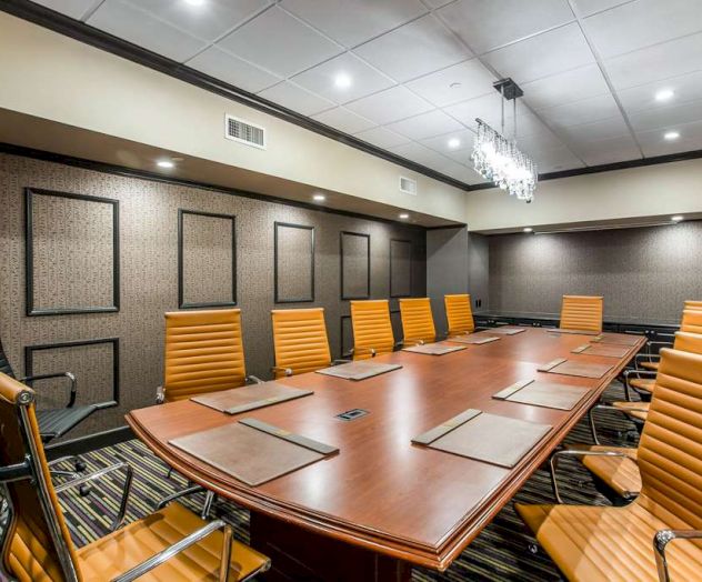 A modern conference room with a large wooden table, orange leather chairs, and a chandelier. Walls are decorated with framed panels.