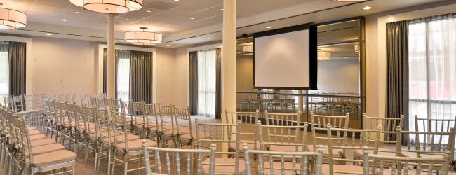 The image shows a conference room with rows of chairs, a screen, and chandeliers, set up for a presentation or event.