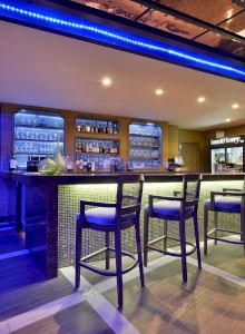 A modern bar with high stools, blue accent lighting, stocked liquor shelves, and a TV on the wall, creating a stylish ambiance.