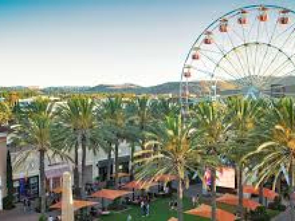 The image shows an outdoor shopping area with palm trees, a Ferris wheel, and people enjoying the space on a sunny day.